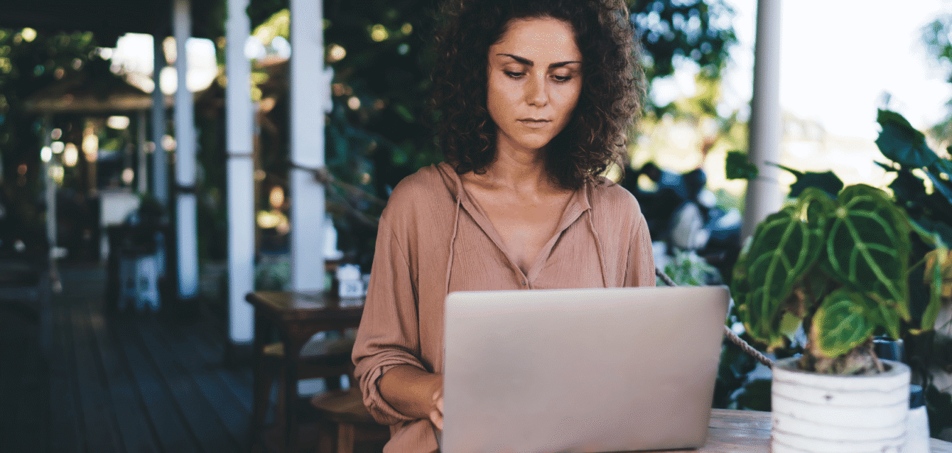 A woman working on her laptop in an outdoor café, representing how Airbnb hosts can leverage referral programs to grow their direct booking network effortlessly Woman with curly hair working on a laptop outdoors at a café, surrounded by plants.